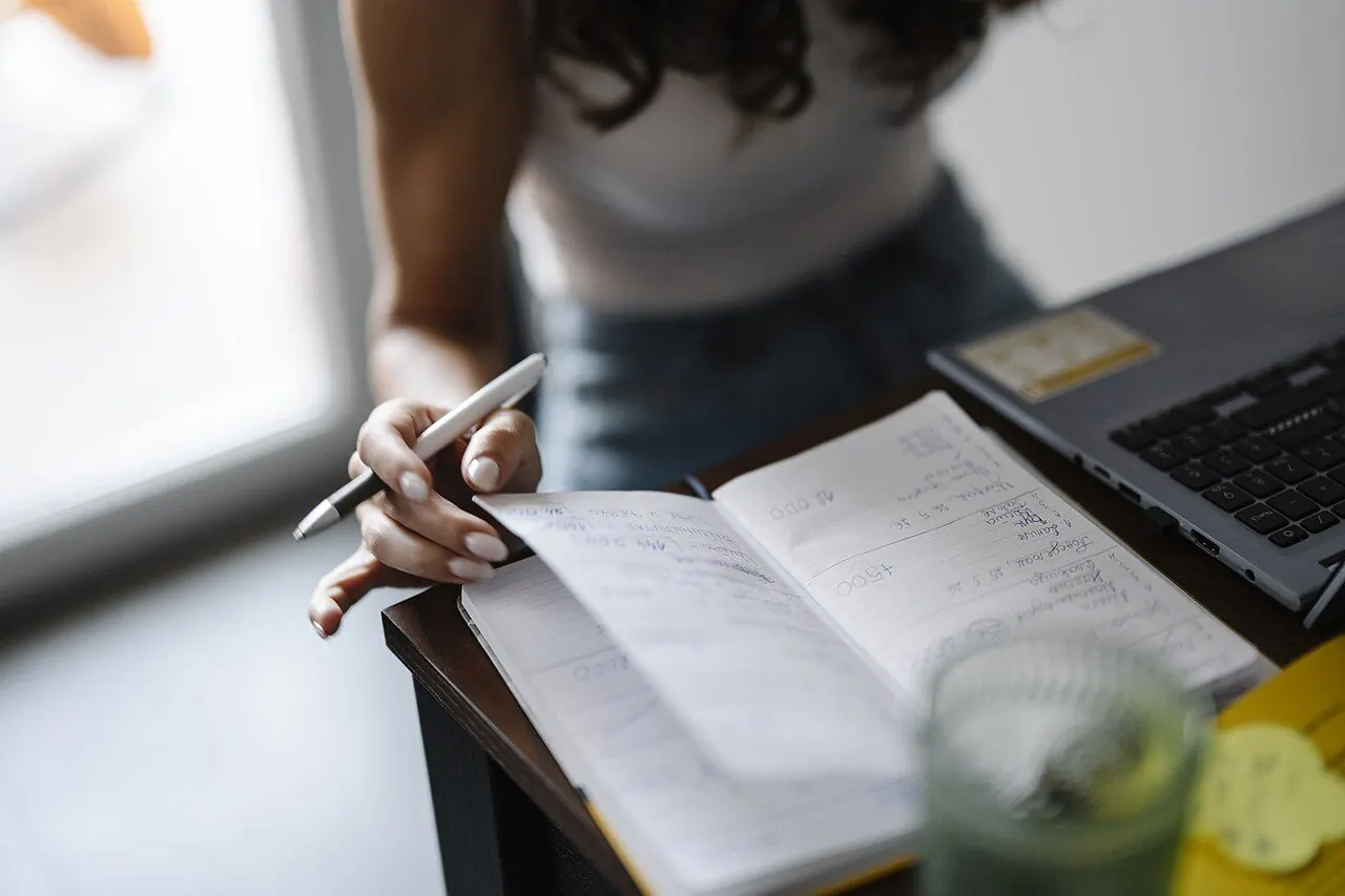 A person is seated at a desk with a laptop open in front. They hold a pen in one hand and flip through a notebook with notes. A glass of water is nearby. Sunlight is coming through a window.
